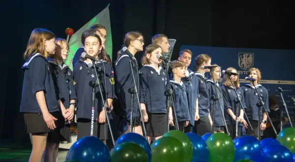 Dzieci w mundurkach śpiewają na scenie, przed nimi mikrofony i balony.