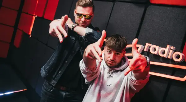 Dwóch mężczyzn pozuje na tle studia radiowego z logo "Zet Radio", wskazując palcami w stronę widza.