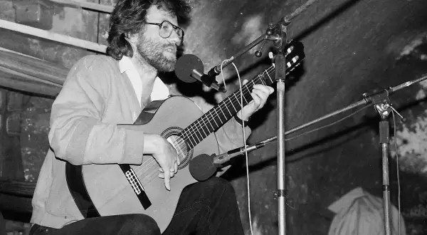 Mężczyzna w okularach gra na gitarze przy mikrofonach.