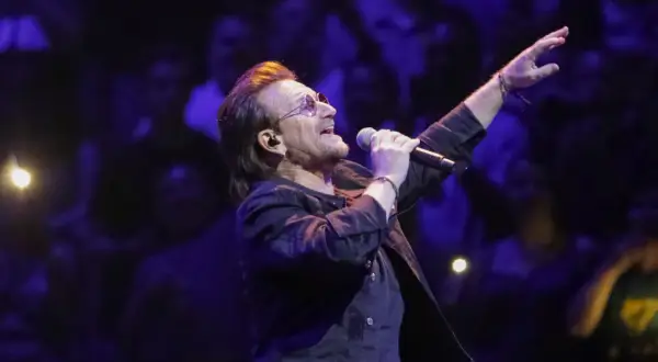 Bono z U2 śpiewa na koncercie, zapowiadając u2 nowa płyta. W tłumie widać światła i sylwetki fanów.