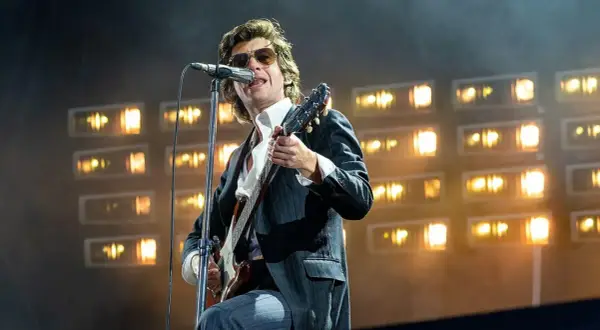 Alex Turner z Arctic Monkeys na scenie, w okularach i garniturze, gra na gitarze podczas jednego z niezapomnianych koncertów.