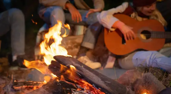 Przy ognisku siedzą ludzie, grają na gitarze i śpiewają piosenki na ognisko.