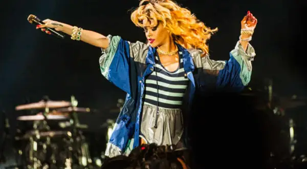 Rihanna na koncercie w Polsce, w bluzie z kolorowymi pasami, śpiewa z mikrofonem w dłoni.