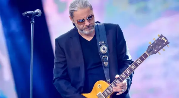 Dżem wokalista z gitarą Gibson na scenie, w okularach przeciwsłonecznych, z siwymi włosami.