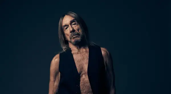 Iggy Pop w czarnej kamizelce, z długimi włosami i zmęczoną miną, na ciemnym tle.