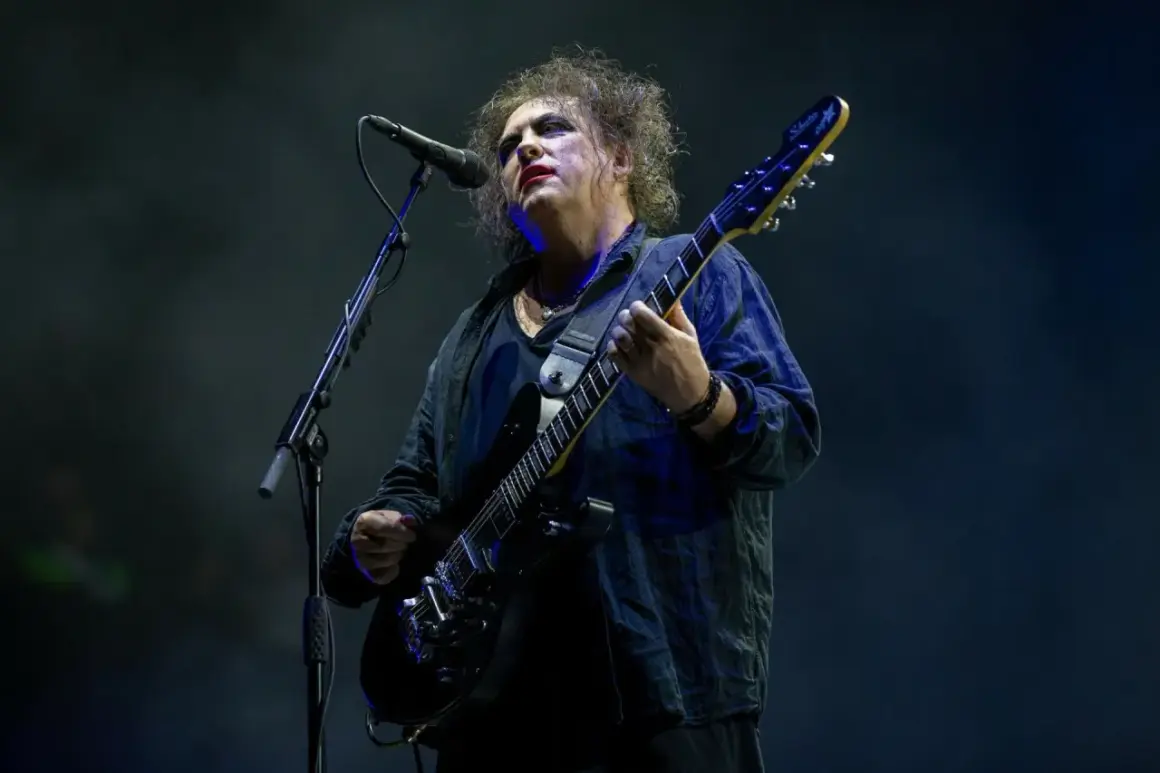 Robert Smith z The Cure na koncercie, w charakterystycznym makijażu, gra na czarnej gitarze elektrycznej.