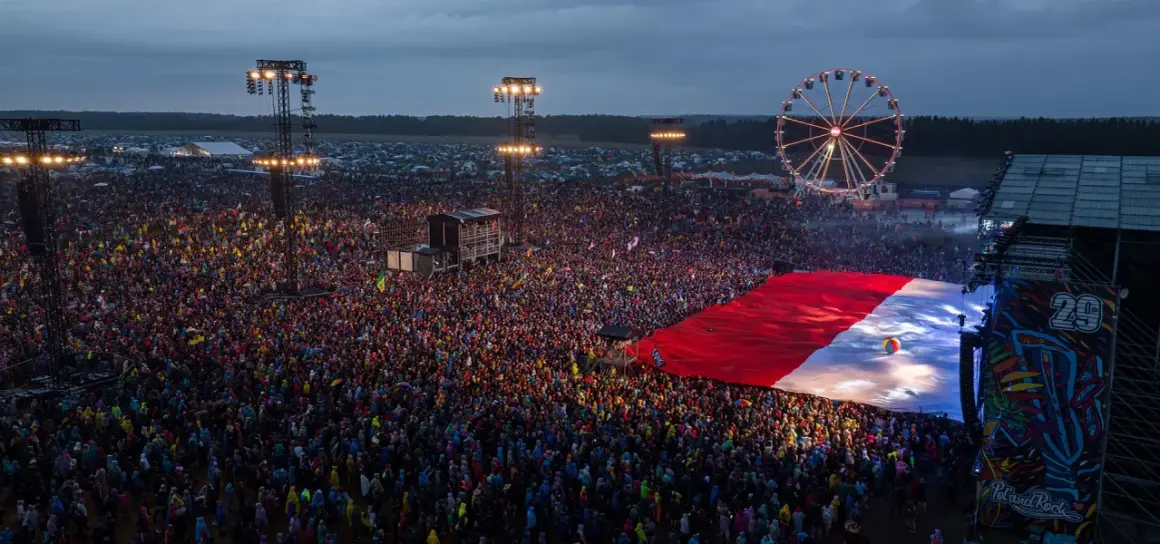 Ogromny tłum ludzi na festiwalu rockowym, rozłożona flaga Polski, scena i diabelski młyn.