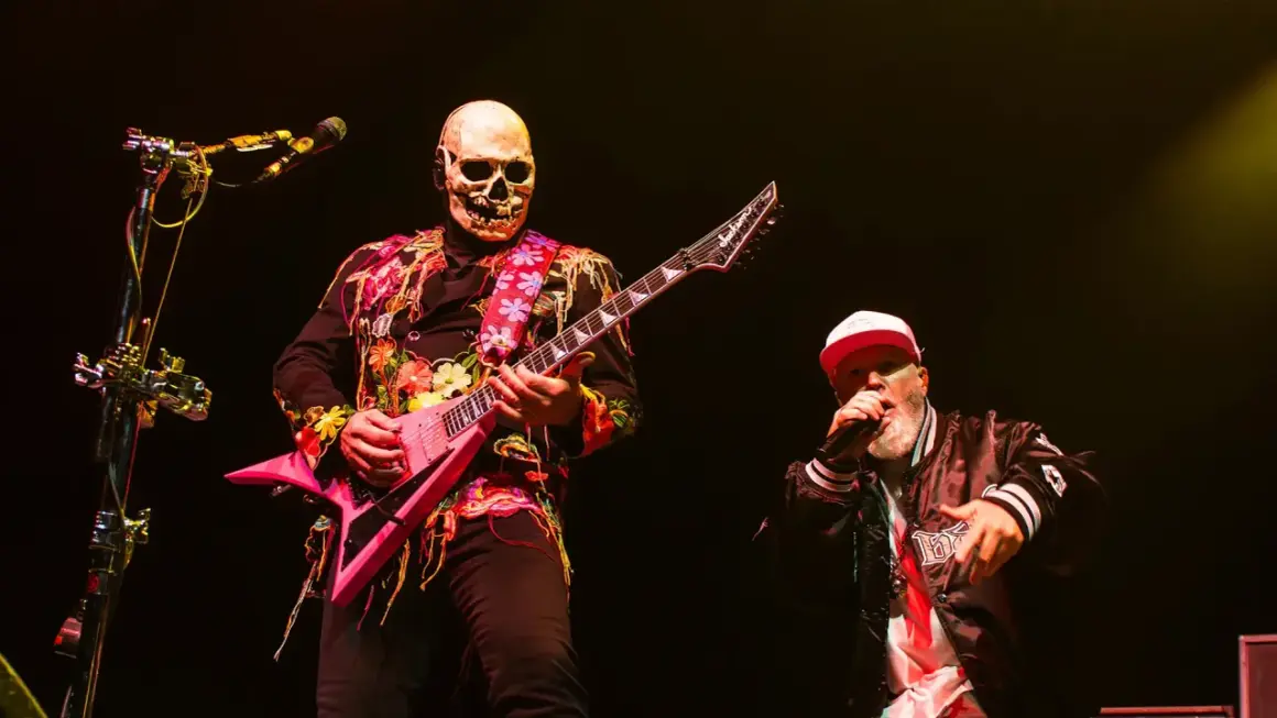 Muzyk w masce czaszki gra na różowej gitarze, a wokalista w czapce śpiewa na koncercie Limp Bizkit.