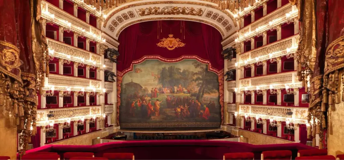 Wnętrze luksusowego teatru operowego z czerwonymi lożami, złotymi zdobieniami i malowidłem na scenie.