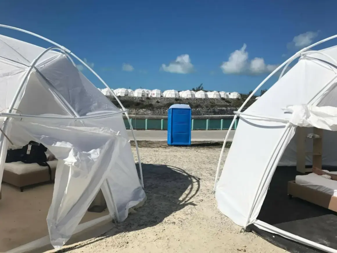 Namioty i niebieska toaleta polowa na plaży, przypominające fiasko Fyre Festival 2. W tle rzędy białych kopuł.