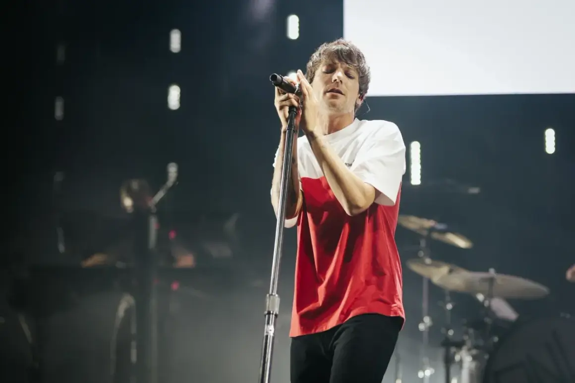 Louis Tomlinson śpiewa na koncercie w Polsce, w czerwono-białej koszulce, z zamkniętymi oczami.