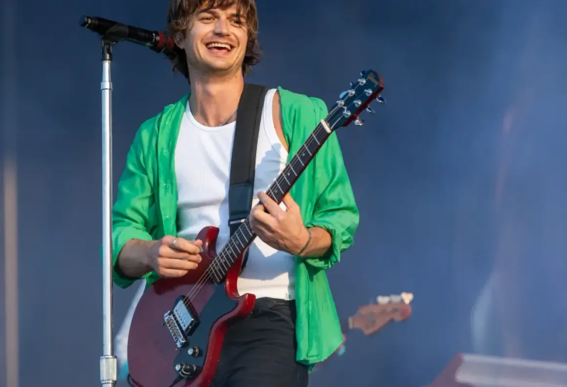 Joe Keery w zielonej koszuli i białej podkoszulce gra na czerwonej gitarze elektrycznej na koncercie.