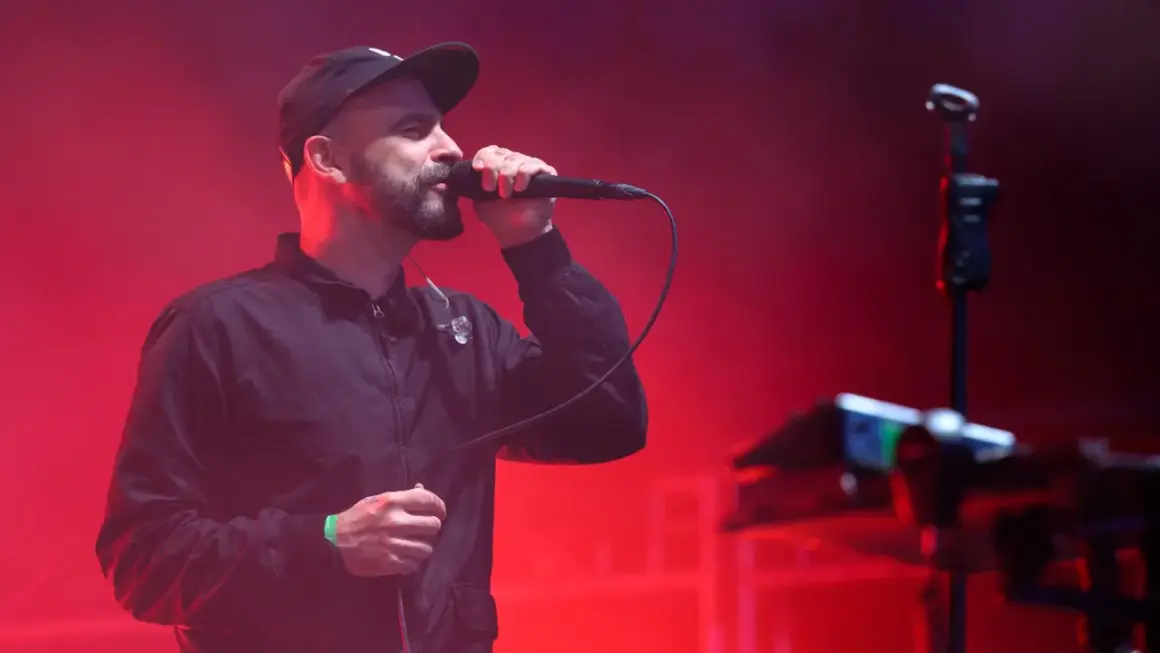 Wokalista z brodą w czapce i kurtce śpiewa do mikrofonu na tle czerwonych świateł. To część tauron nowa muzyka line-up.