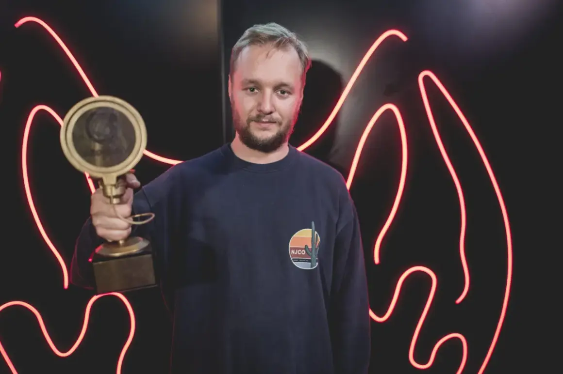 Mężczyzna w granatowej bluzie trzyma złotą statuetkę na tle czerwonych neonów.
