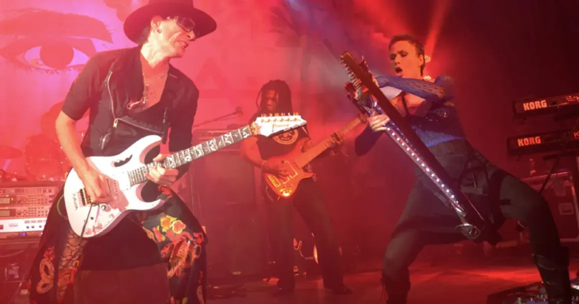 Trzech gitarzystów na scenie, jeden w kapeluszu gra na białej gitarze, drugi w tle na pomarańczowej, trzeci na futurystycznej.