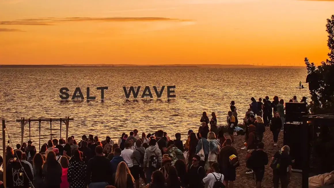 Ludzie na plaży podczas zachodu słońca, z napisem "SALT WAVE" na wodzie.
