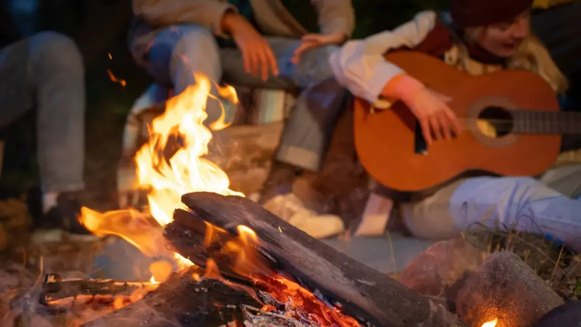 Przy ognisku siedzą ludzie, grają na gitarze i śpiewają piosenki na ognisko.