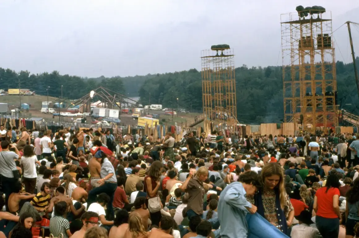 Tłum ludzi na festiwalu Woodstock, z widocznymi wieżami i sceną w tle.