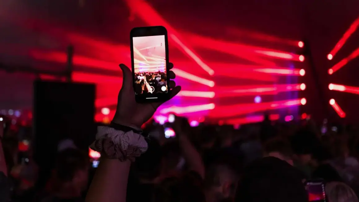 Dłoń trzyma smartfon nagrywający koncert z czerwonymi światłami w tle.