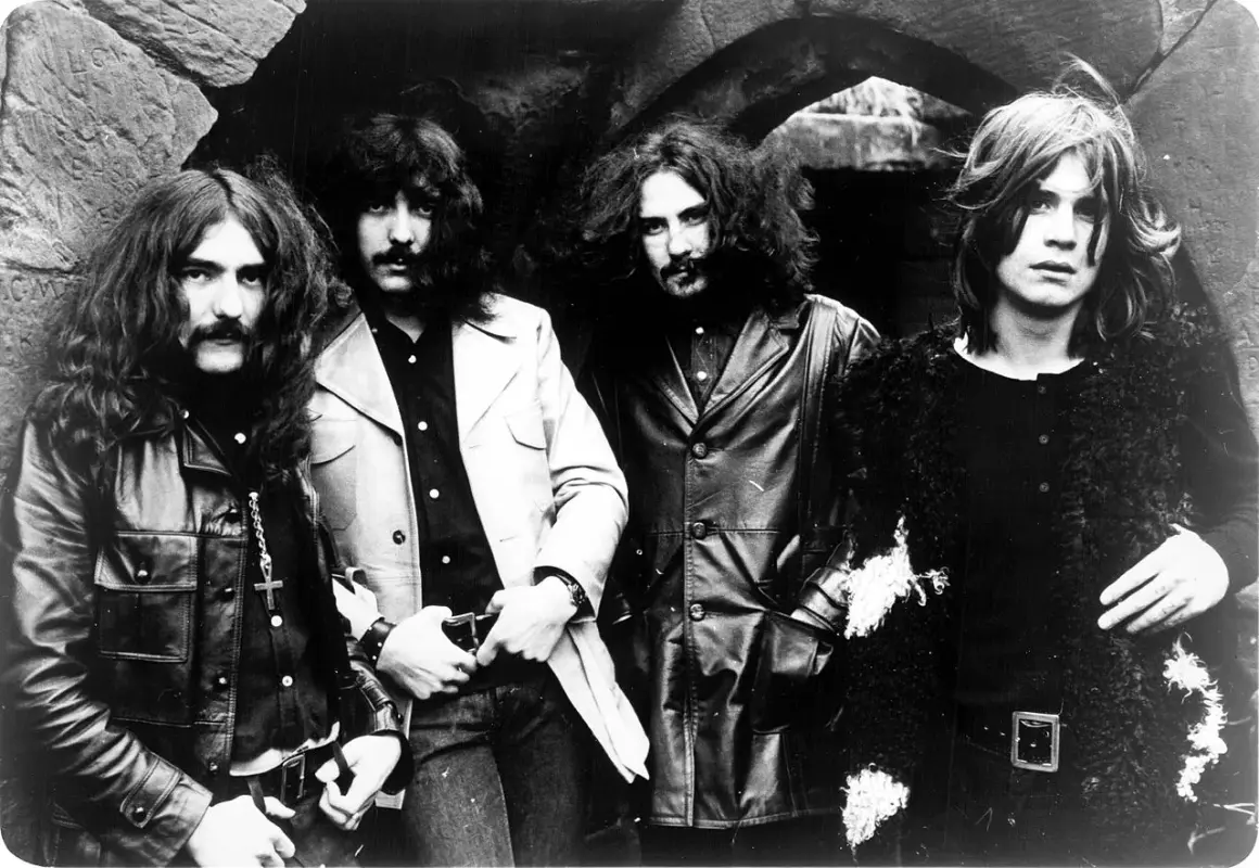 Czterech członków zespołu Black Sabbath pozuje do zdjęcia w mrocznym, kamiennym otoczeniu.