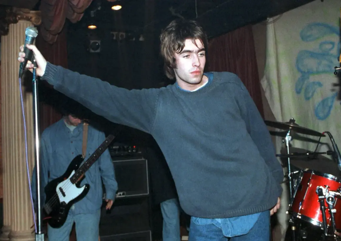 Liam Gallagher z Oasis śpiewa na scenie, w tle basista i perkusja. To klasyczne oasis utwory.
