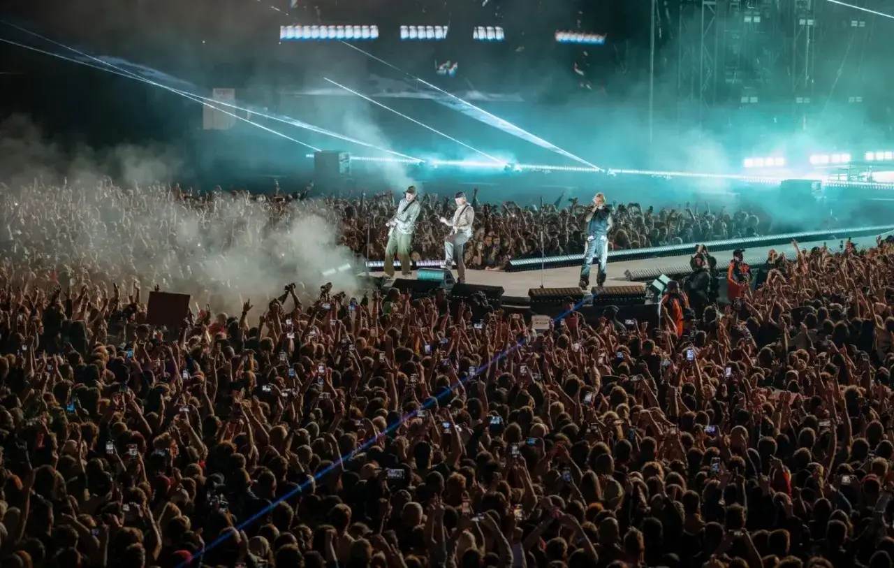 Tłum szaleje podczas koncertu Linkin Park. Na scenie trzej muzycy, a przed nimi morze rąk z telefonami.