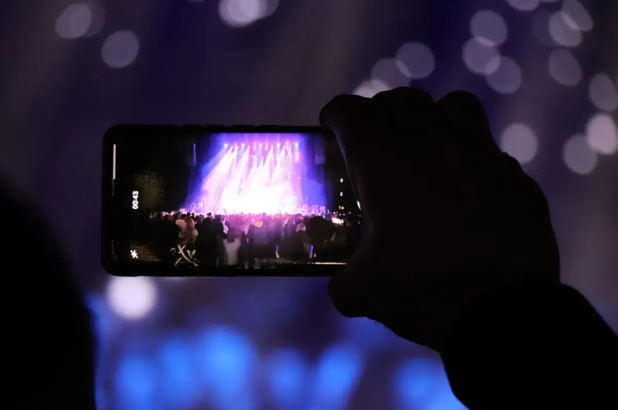 Ręka trzyma telefon, nagrywając koncert na krakowskim festiwalu muzycznym. Na ekranie widać scenę oświetloną kolorowymi światłami i tłum bawiących się ludzi.