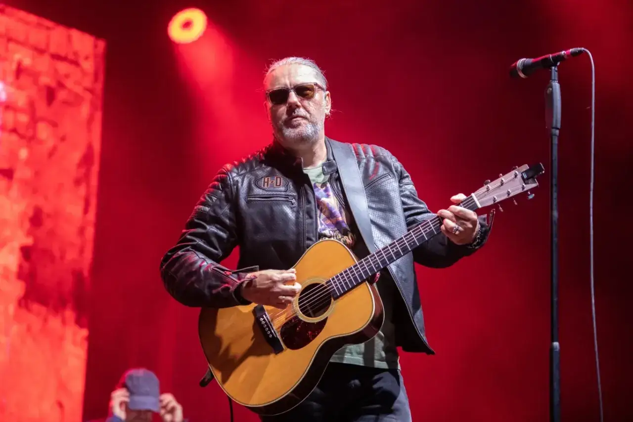 Dżem wokalista z gitarą na scenie, w skórzanej kurtce i okularach przeciwsłonecznych.