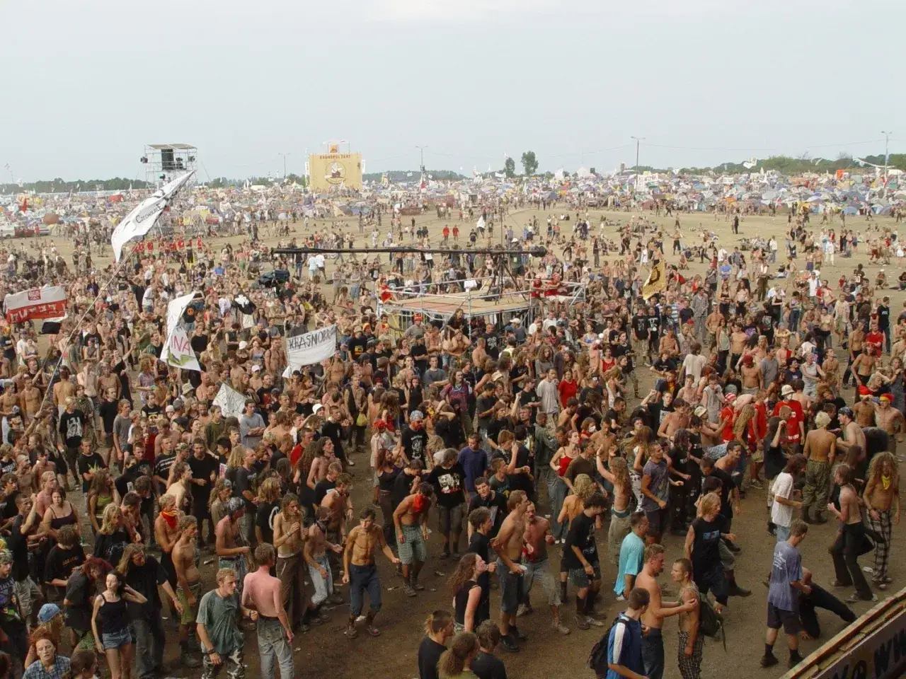 Tłum ludzi na festiwalu, gdzie jest Woodstock. Widać scenę, namioty i flagi.
