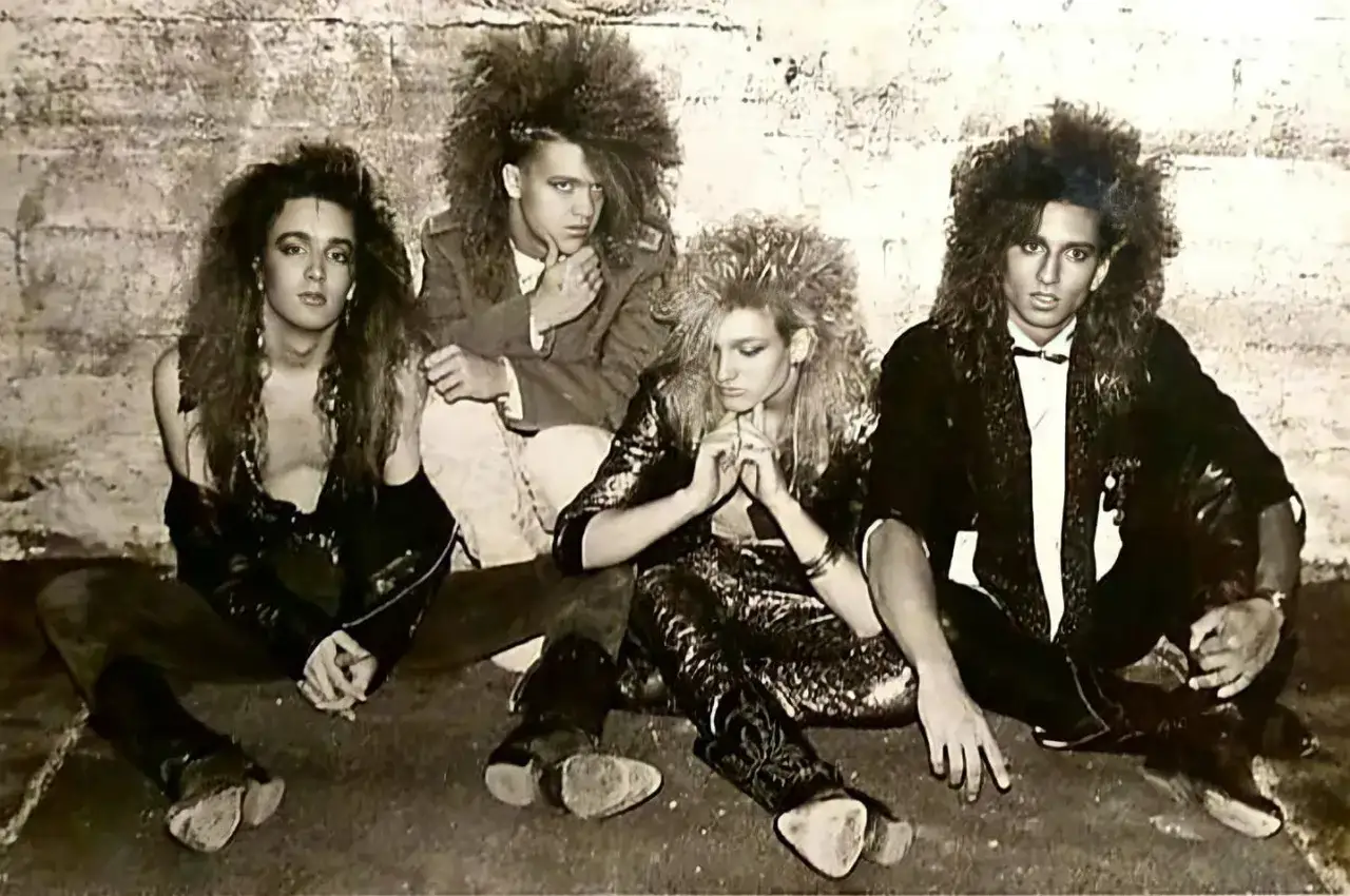Członkowie Alice in Chains z charakterystycznymi fryzurami i strojami z lat 80. siedzą na ziemi przed ceglaną ścianą.