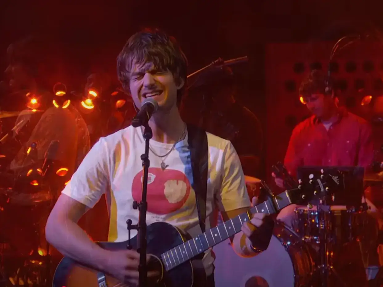 Joe Keery śpiewa i gra na gitarze na scenie, ubrany w białą koszulkę z nadrukiem jabłka.