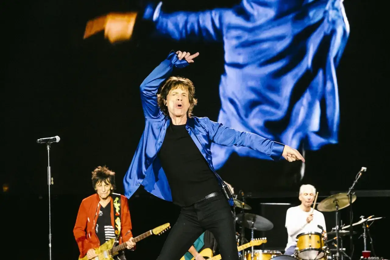 Mick Jagger w niebieskiej koszuli tańczy na scenie podczas jednego z koncertów The Rolling Stones. Obok Ronnie Wood gra na gitarze, a Charlie Watts przy perkusji.
