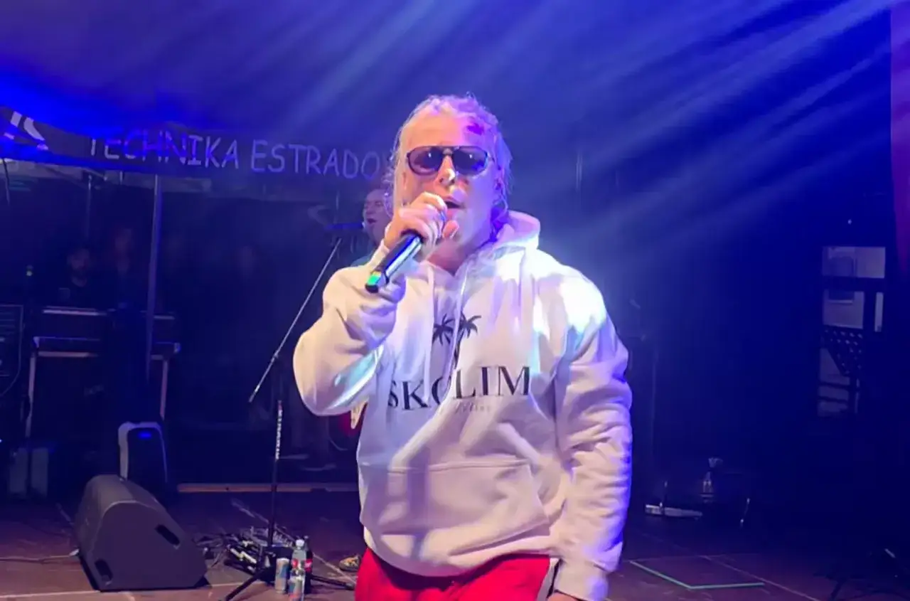 Artysta w białej bluzie z kapturem i okularach przeciwsłonecznych śpiewa z playbacku na scenie, oświetlony niebieskim światłem.