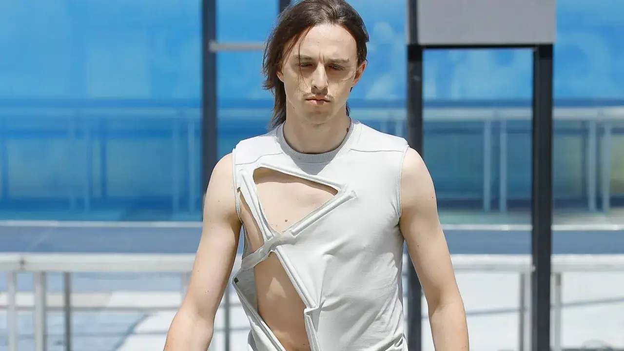 Tommy Cash w szarej, podartej koszulce bez rękawów, prezentuje odważny styl na tle błękitnego budynku.