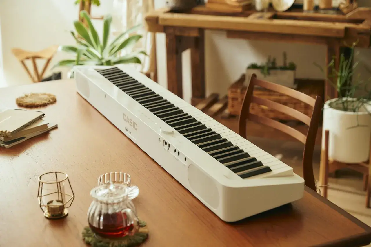 Biały keyboard Casio leży na drewnianym stole. Obok stoją ozdobne świeczniki i dzbanek z herbatą.
