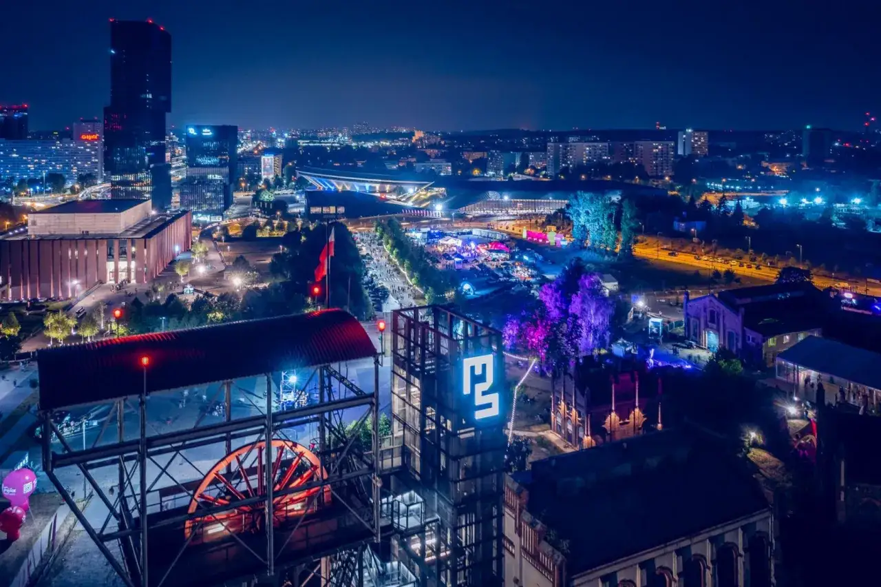 Nocny festiwal Tauron Nowa Muzyka w Katowicach. Widać festiwalowy teren z tłumami, scenami i iluminacjami, a w tle panoramę miasta.