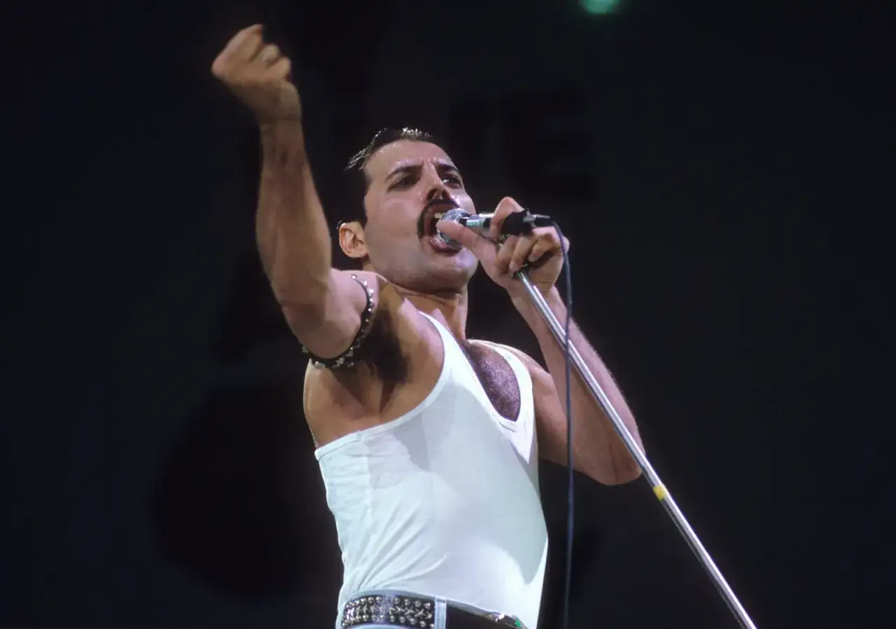 Freddie Mercury w białej podkoszulce, z uniesioną pięścią, śpiewa na jednym z niezapomnianych Queen koncerty.