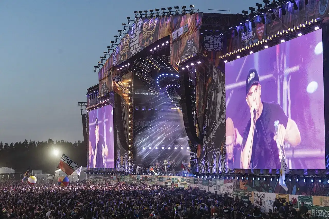 Koncert na Pol'and'Rock Festival, dawniej Przystanek Woodstock. Tłum ludzi, wielkie ekrany z wokalistą, scena z oświetleniem.