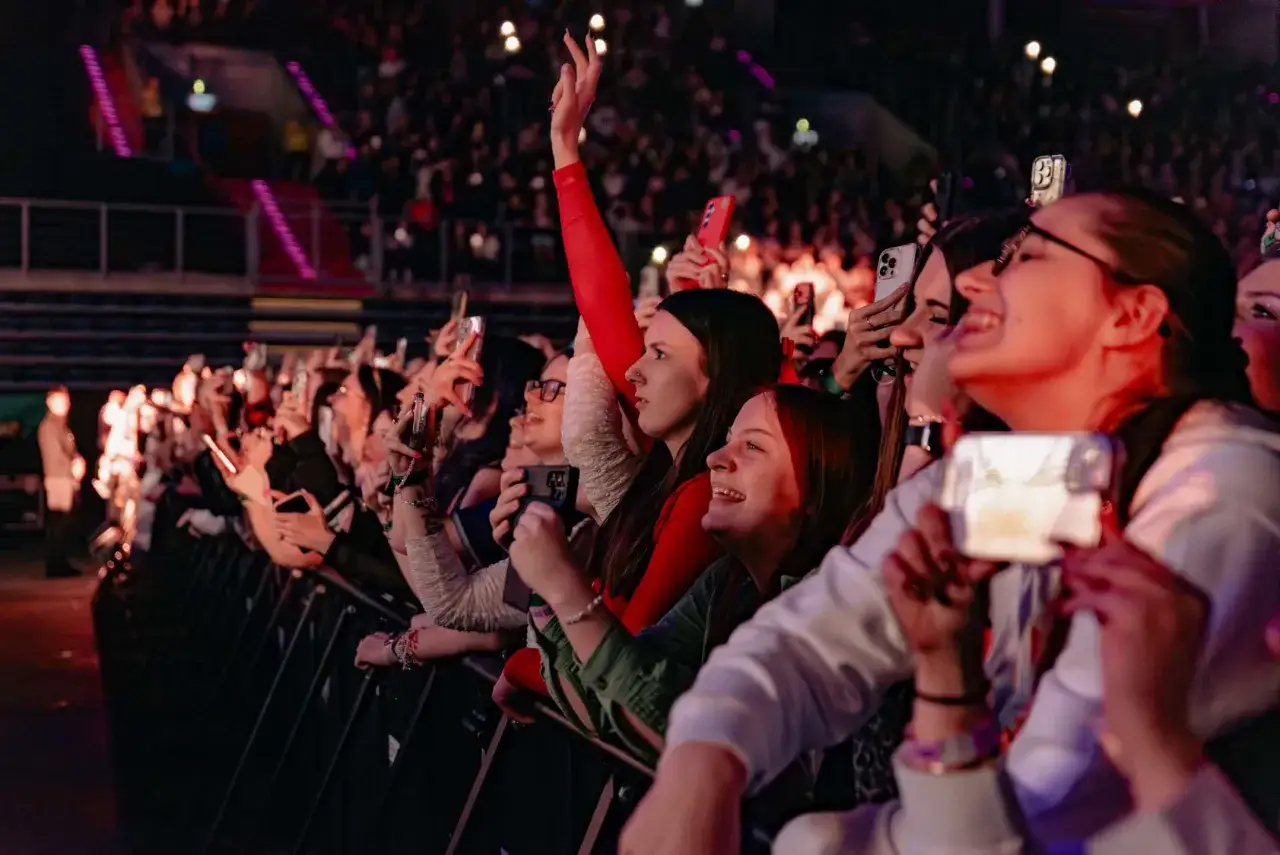 Uśmiechnięci fani z telefonami w dłoniach, nagrywający koncert. Widać fragmenty sceny i tłum.