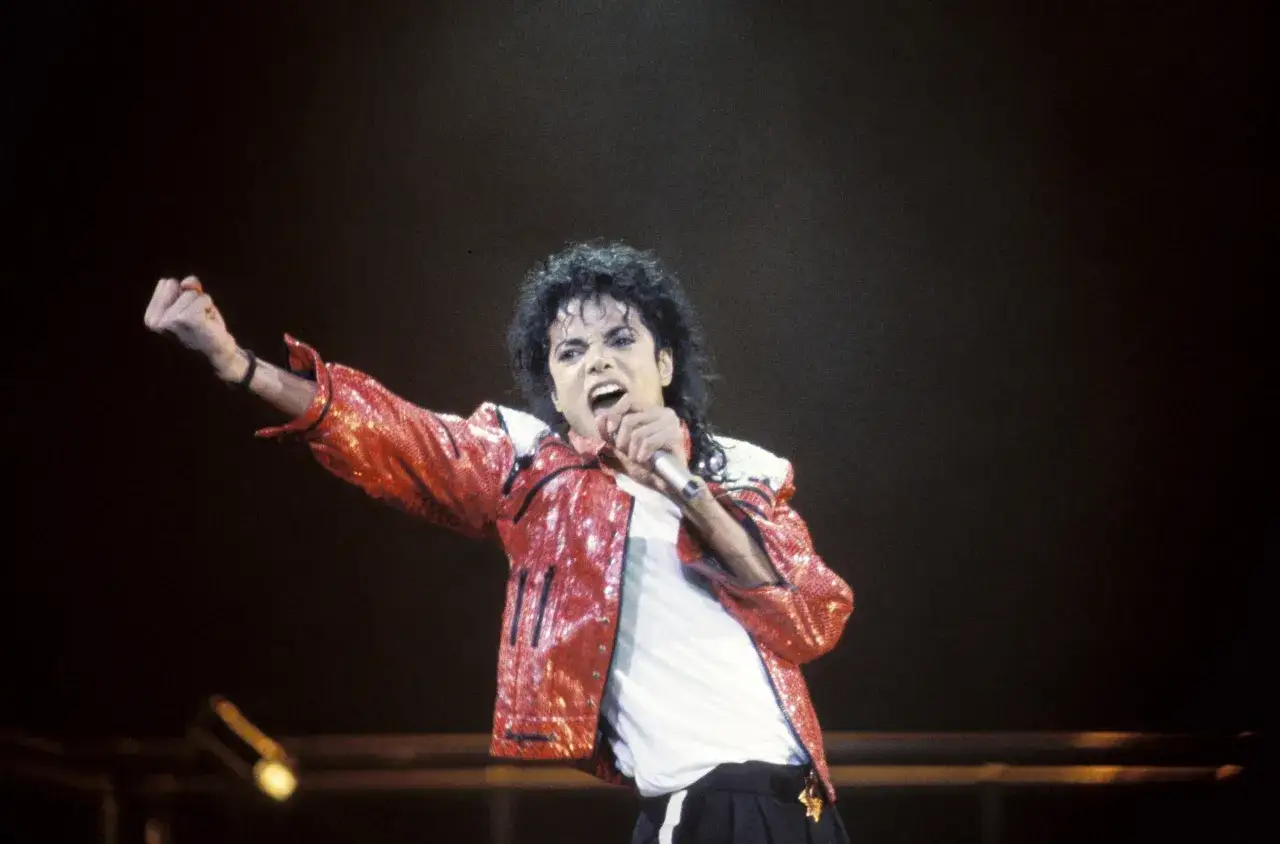 Król popu, Michael Jackson, w błyszczącej czerwonej kurtce śpiewa na scenie, unosząc pięść w geście triumfu.