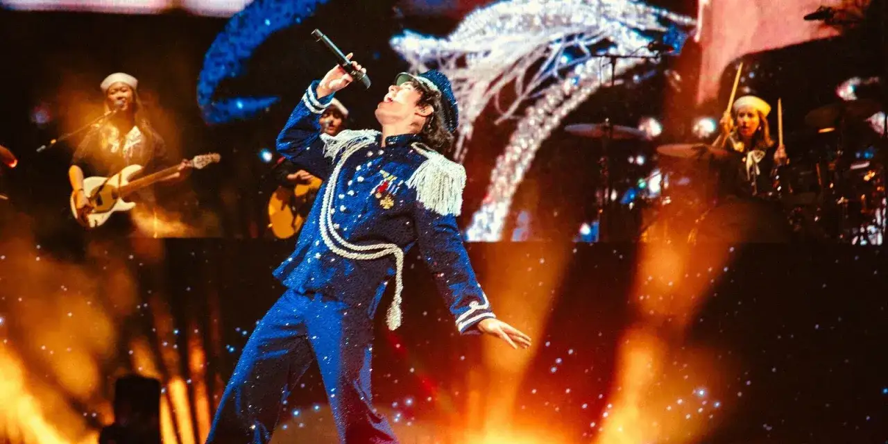 Conan Gray w błyszczącym, granatowym stroju z frędzlami śpiewa na koncercie. Na scenie grają też gitarzystka i perkusistka.