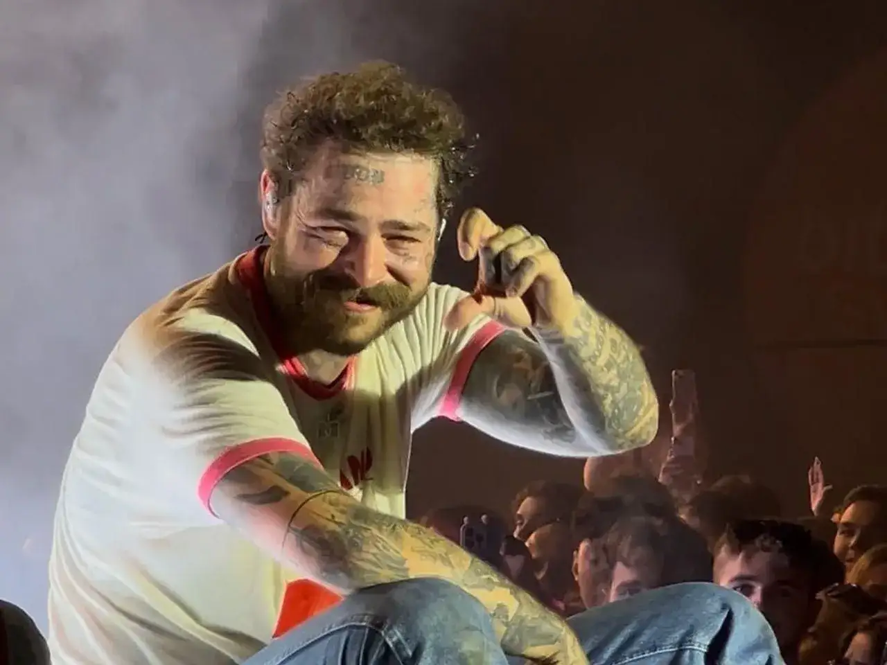 Post Malone na koncercie, uśmiechnięty, z tatuażami na twarzy i rękach, pozdrawia tłum.