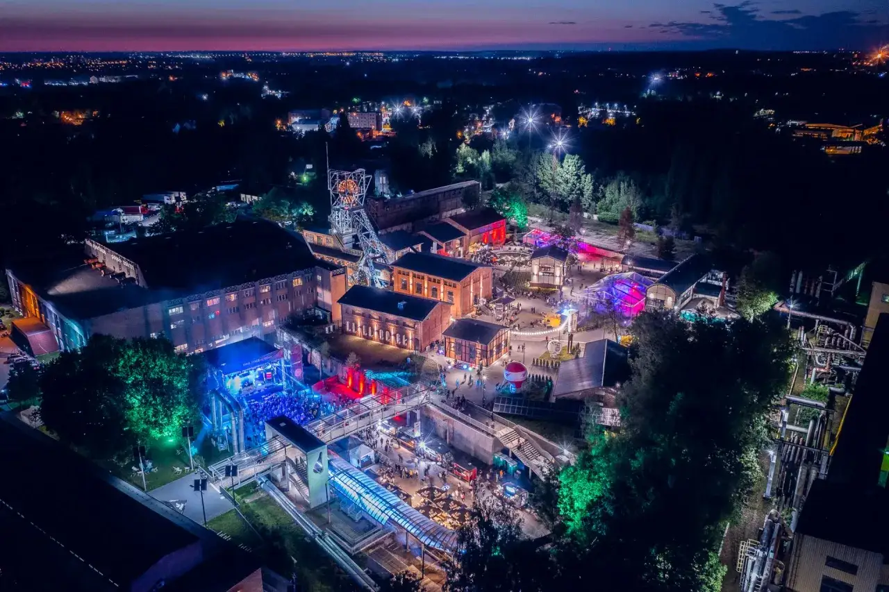 Nocny festiwal w industrialnej scenerii, rozświetlony kolorowymi światłami. Widać scenę z tłumem, budynki kopalni i drzewa. To miejsce tętni życiem podczas carbon festival.