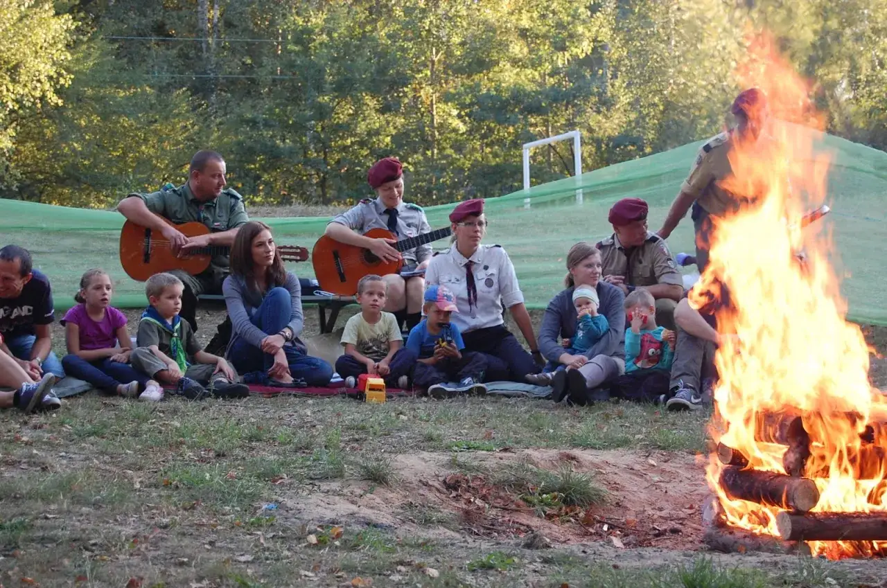 Grupa harcerzy i dzieci siedzi przy ognisku, słuchając piosenek harcerskich granych na gitarach.