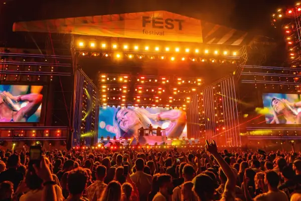 Tłum na festiwalu, scena z wielkim ekranem i logo "FEST festival program". Światła i artyści na scenie.