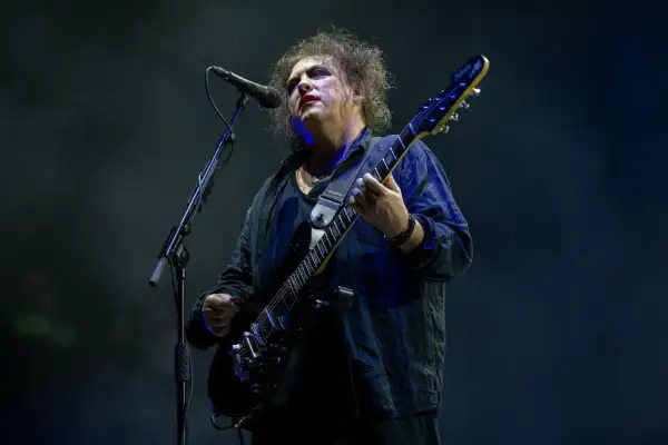 Robert Smith z The Cure na koncercie, w charakterystycznym makijażu, gra na czarnej gitarze elektrycznej.