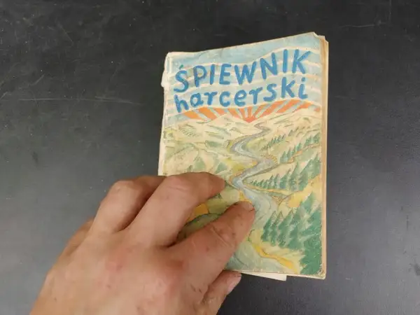 Dłoń trzyma stary śpiewnik harcerski z rysunkiem rzeki i gór.