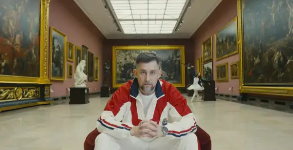 Mężczyzna w dresie siedzi w galerii sztuki, otoczony obrazami i rzeźbami.