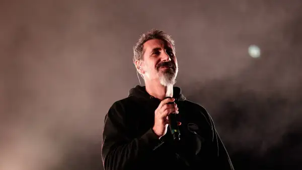 Serj Tankian z System of a Down na koncercie, trzyma mikrofon, w tle mgła i światło.