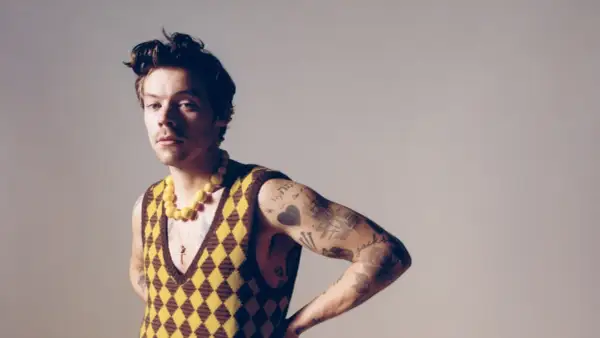 Harry Styles w kamizelce w romby i żółtym naszyjniku, z tatuażami na ramieniu.